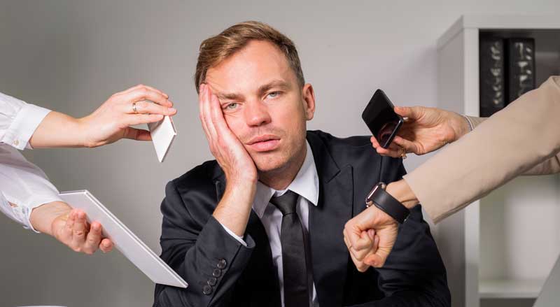 A tired businessman at his desk with two people holding a phone in his face and showing him the time on their watch