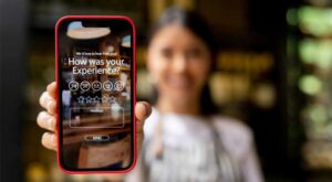 A woman holding a smartphone toward the camera displaying a customer feedback screen asking, “How was your experience?”